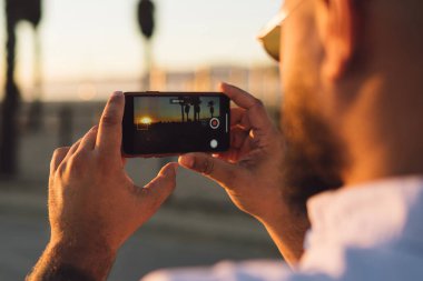Gün batımı sırasında tatil, modern smartphone ve uygulama alarak resim Doğa gezisi sırasında kullanılarak hipster adam güzel manzara çekim için erkek holding cep telefonunuzun arka görünümü kadar kapatın