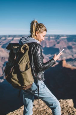 Gülümseyen kadın Amerikalı turist smartphone cep telefonu ile iyi veri bağlantısında dağlar, sırt çantası elinde vahşi doğa'da cliff yürüyüş üzerinde duran cep bakarak kadınla memnun mesajı okumak