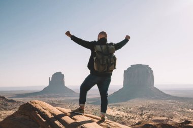 Sırt çantalı erkek gezgin, Anıt Vadisi 'nin güzel doğasıyla heyecanlanıyor. Yüksekteki kayaların üzerinde duruyor. Elleri havada, hipster kız Arizona' ya yaptığı başarılı yolculuktan dolayı mutlu.
