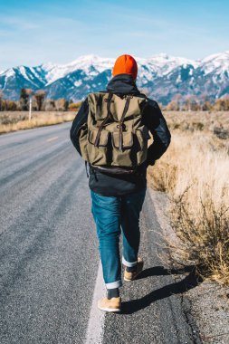 Kış, dağ tepeleri karlı zirveleri ile yürüyerek otostop yolculuk sırasında almak sırt çantası ile hipster adam, vahşi çevre keşfetmek için asfalt yol oluyor yürüyüş erkek Gezgin görünümünü geri