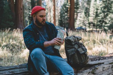 Erkek yolcu Yolculuk tutkusu dinlenmek doğa ortamı, ABD'de yolculuk sırasında vahşi orman keşfetmek sırt çantası ile eski ağaçta oturuyor hipster adam keşfetmek haritada rota kontrol ahşap doğa yürüyüşü