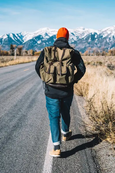 Kış, dağ tepeleri karlı zirveleri ile yürüyerek otostop yolculuk sırasında almak sırt çantası ile hipster adam, vahşi çevre keşfetmek için asfalt yol oluyor yürüyüş erkek Gezgin görünümünü geri