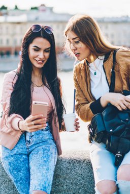 İki neşeli arkadaşlarım şık siyah sırt çantası ve lezzetli kahve el modern telefon şehir promosyon arka plan üzerinde oturan yüklü yeni fotoğraf uygulaması ile selfie resim yapma