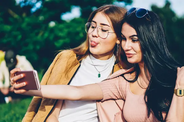 Arkadaşlarım şık modern smartphone aygıt Park ön kamera komik selfies yapma. Esmer ve sarışın hippi kız açık havada oturup telefon üzerinde fotoğraf çekimi birlikte eğleniyor