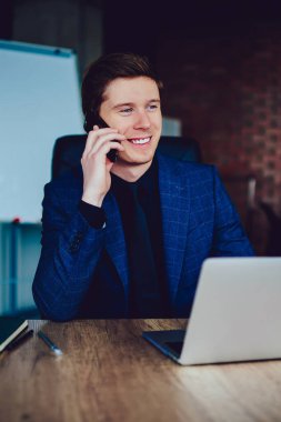 Gülümseyen erkek yönetici müşteri hizmetlerine hesap bakiyesini kontrol etmeleri için sesleniyor. Haberlerden heyecanlanan başarılı iş adamı telefon görüşmeleri sırasında netbook ile ahşap masada oturuyor.