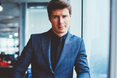 Kendine güvenen şık takım elbiseli bir işadamının portresi ofis penceresinin yanında duruyor ve kameraya bakıyor, yakışıklı beyaz erkek girişimci çalışma alanında vakit geçiriyor ve iş arkadaşını bekliyor.