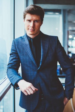 Kendine güvenen şık takım elbiseli bir işadamının portresi ofis penceresinin yanında duruyor ve kameraya bakıyor, yakışıklı beyaz erkek girişimci çalışma alanında vakit geçiriyor ve iş arkadaşını bekliyor.