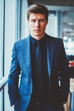 Zarif takım elbiseli ciddi uzman ekonomistin yarı boy portresi ceplerinde, modern şirket ofisinde dikilirken resmi giyinmiş şık, kendine güvenen iş adamı kameraya bakıyor.