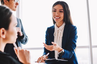 Esmer saçlı, pozitif gülümseyen kadın iş arkadaşı profesyonel iş arkadaşlarına, başarılı iş adamı ve kadın takımının modern ofis stratejisi hakkında konuştuklarını anlatıyor.