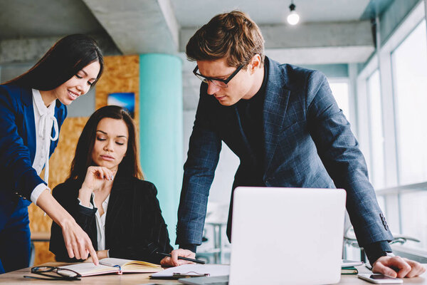 Young business team discussing strategy ideas near desktop while reading information from textbook, confident male and female professionals consultancy to each other searching solving for problem