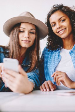 İki mutlu çok kültürlü bayan arkadaş birlikte vakit geçirip içeride eğleniyorlar. Modayı takip eden hippi kız modern akıllı telefon cihazından, yanında oturan kadına bilgi aktarıyor.