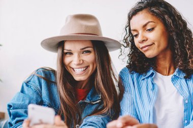 Neşeli kadın blog yazarının portresi telefon cihazıyla fıkra okumaktan ve kameraya bakmaktan gülerken yanında oturan ve akıllı telefon dokunmatik ekranına bakan kadın arkadaşının portresi.