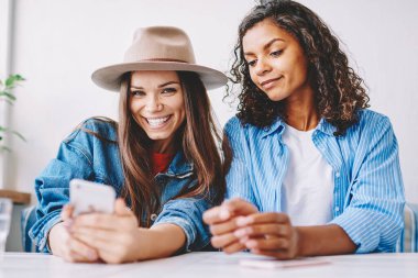 Neşeli kadın blog yazarının portresi telefon cihazıyla fıkra okumaktan ve kameraya bakmaktan gülerken yanında oturan ve akıllı telefon dokunmatik ekranına bakan kadın arkadaşının portresi.