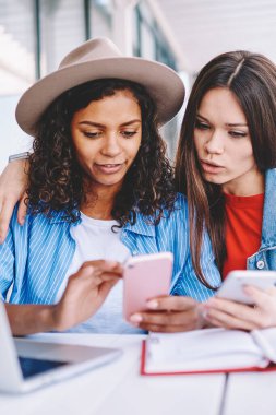 Akıllı telefon cihazları hakkında bilgi paylaşan çekici kadınlar dizüstü bilgisayarla aynı masada oturuyorlar iyi kablosuz bağlantı kullanıyorlar, çok kültürlü hipster kızları cep telefonuyla takipçilerle mesajlaşıyorlar.