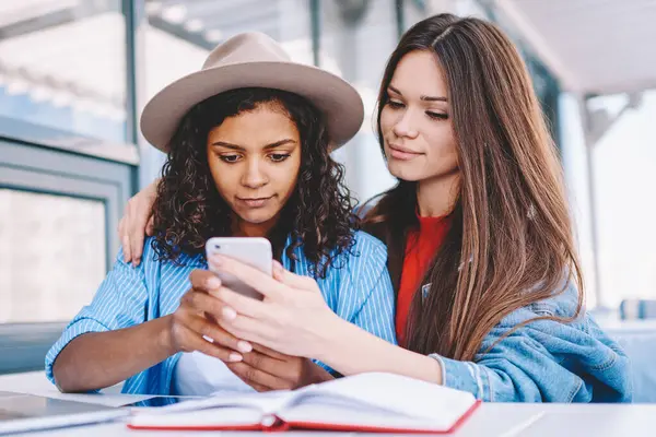 Gündelik giyinmiş çekici hippi kız, yanında oturan ve dijital akıllı telefon cihazıyla e-posta alan kız arkadaşına sarılıyor, cep telefonuyla multimedya uygulaması üzerinden video izleyen büyüleyici kadınlar.