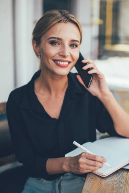 Neşeli kız öğrencinin portresi dışarıda dizüstü bilgisayar ve eğitim kitaplarıyla oturmuş akıllı telefon ve gülümsemeyle başarılı bir şekilde iletişim kuruyor.