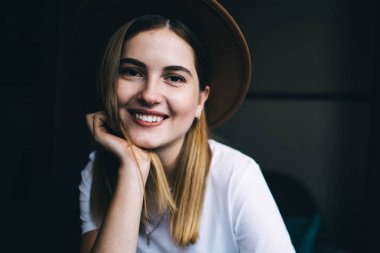 Moda şapkalı, siyah arka planda oturan neşeli 20 'li yaşlardaki hippi kızın yarım boy portresi. Gülümsüyor, şık giyinmiş, kameraya bakarak mutlu, genç bir kadın.