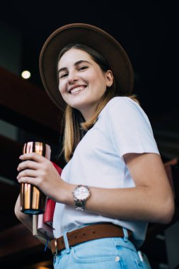 Şapkalı, şapkalı, gülümseyen, boş zamanın tadını çıkaran neşeli beyaz kadın portresi kitap ve içecekle termo bardak taşıyan zengin genç hippi kız.