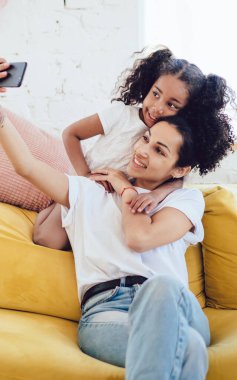 Siyah gülümseyen genç bir kadın neşeli kıvırcık kızıyla selfie çekiyor. Sarı kanepede arkasında büyük bir yastıkla oturuyor.