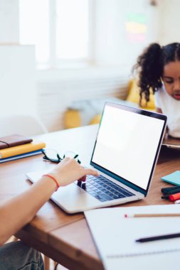 Modern evde tablet kullanan, beyaz ekranlı, küçük kızıyla aynı masada oturan, cep telefonuyla konuşan, serbest çalışan genç bir kadının yan görüntüsü.