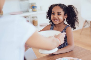 Masanın yanında şekerli tanınmayan bir kadın büyük oda arka planında kurabiye pişirirken dağınık suratlı, gülümseyen, kıvırcık, etnik bir kıza kase veriyor.