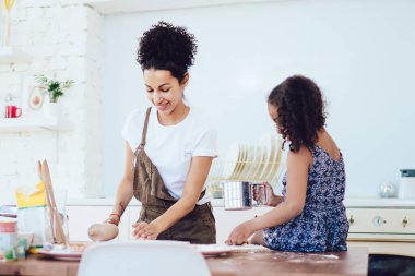 Genç, neşeli siyah anne mutfak masasında gülümseyip hamuru yuvarlarken kıvırcık saçlı kız elek ve un ellerken