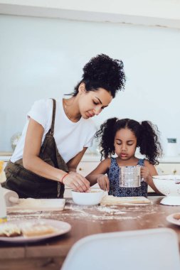 Afro-Amerikalı kıvırcık kadın ve kızı kurabiye yaptığı için un seçiyor ve evdeki büyük açık mutfağa kase koyuyor.