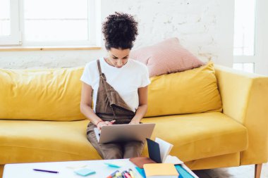 Kıvırcık saçlı, serbest giyinmiş, sarı koltukta otururken dizüstü bilgisayarda çalışan Afro-Amerikalı bir kadın.