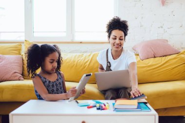 Neşeli Afro-Amerikalı kadın dizüstü bilgisayarı ile parlak odada oturuyor ve masadan defter alırken tabletli kıvırcık kız yanında oynuyordu.