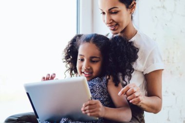 Beyaz tişörtlü Afro-Amerikan esmer anne evde giydiği güzel kıvırcık kızıyla oturuyor ve pencerenin yanında tablet kullanıyor.
