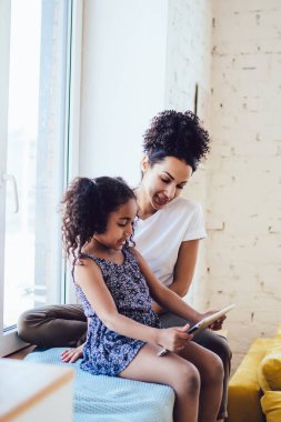 Beyaz tişörtlü kıvırcık Afrikalı Amerikalı annenin, esmer kızıyla pencere eşiğinde oturup, evde birlikte tablet kullanışının yan görüntüsü.
