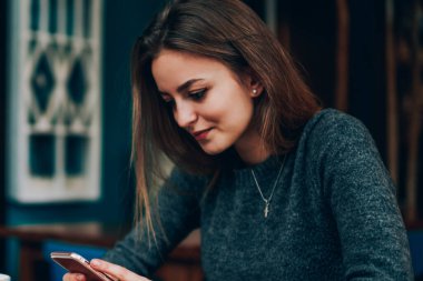 Kablosuz internete bağlı modern elektronik cihazla Kafkasyalı kız öğrenci çevirme numarası pozitif, güzel hippi kız kafeteryada eğleniyor telefon ağlarındaki yeni güncellemeleri kontrol ediyor.