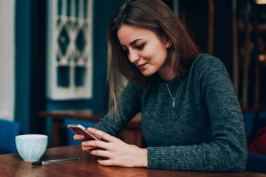 Kablosuz internete bağlı modern elektronik cihazla Kafkasyalı kız öğrenci çevirme numarası pozitif, güzel hippi kız kafeteryada eğleniyor telefon ağlarındaki yeni güncellemeleri kontrol ediyor.