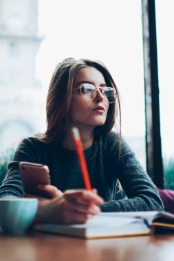 Düşünceli kadın blogcu, kampüs kafeteryasında sosyal ağ ile ilgili ilginç bir fikri olan yeni bir yazı yazmayı planlarken düşünceli bir şekilde kenara bakıyor. Kapalı alanda not defteriyle çalışan düşünceli hippi kız.
