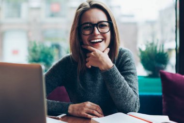 Başarılı gülümseyen yeniyetme kadın portresi serbest çalışmadan, modern dizüstü bilgisayardan, görüş koruması için gözlük takan bayan öğrenciden, kapalı alan kitabı ve netbook kullanarak.