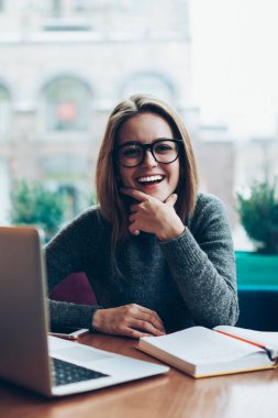 Başarılı gülümseyen yeniyetme kadın portresi serbest çalışmadan, modern dizüstü bilgisayardan, görüş koruması için gözlük takan bayan öğrenciden, kapalı alan kitabı ve netbook kullanarak.