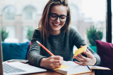 Moda gözlüklü pozitif bayan öğrenci çalışma alanında modern defterle eğleniyor, genç, hippi kız blog kitabı için kafeteryayı ziyaret ettiğine dair makale eleştirisi yazıyor.