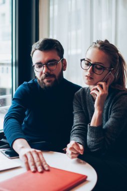 Yoğunlaşmış iki arkadaş birlikte çalıştığı yerde dizüstü bilgisayarla iş birliği yapıyor. Gözlüklü ciddi bir kadın, iş arkadaşının yanında otururken akıllı telefondan önemli bir konuşma yapıyor.