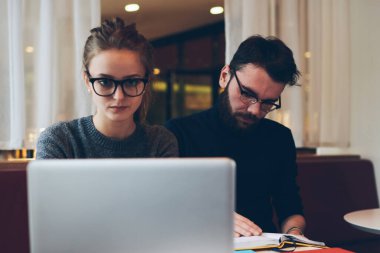 Moda gözlüklü, görüş düzeltmesi için odaklanmış bir kadın kafeteryadaki kablosuz internete bağlı dizüstü bilgisayarda internetten web sitesi izlerken, düşünceli erkek arkadaş ders kitabında bilgi arıyor.