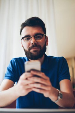 Gözlüklü genç yakışıklı hippi portresi. Mesaj yazmak için modern cep telefonunu kullanıyor ve kameraya bakıyor. Milenyum erkeği ara verdiğimizde sohbet etmek için akıllı telefon tutuyor.