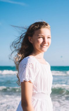 Beyaz elbiseli, gülümseyen sarışın bir kız. Mavi okyanusun kıyısında, uçurtma sörfçüleriyle dikiliyor ve bulutsuz güneşli havayı seyrediyor.