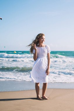 Uçurtma sörfçüleri ile dalgalı deniz kenarında çıplak ayakla dikilen, şık beyaz elbiseli, uzun boylu, gülümseyen bir kız.