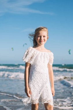 Beyaz dantelli, sarı saçlı, pozitif gülümseyen küçük bir kız güneşli bir günde kumsalda tatilin tadını çıkarıyor.