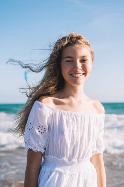 Yazlık elbiseli, pozitif gülümseyen küçük sarışın kumlu sahilde duruyor parlak güneşin altında sallanıyor ve kameraya bakarken gülümsüyor.