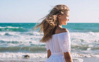 Beyaz elbiseli, düşünceli bir genç kızın kumlu sahilde tek başına dinlenirken dalgaların sesini dinlemesi.