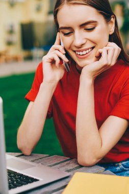 Arkadaşlarıyla akıllı telefondan konuşan ve dışarıda dinlenirken laptopunu kullanan neşeli bayan öğrenci, genç profesyonel serbest çalışan iş görüşmeleri için meslektaşlarını arıyor.