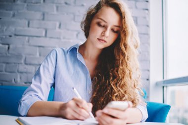 Ciddi hippi kız, okul ödevi yapan çekici kız öğrenci ve ücretsiz 4G internet kullanarak akıllı telefon cihazı üzerinde araştırma yaparken ders kitabına organizasyon planı yazmak için zaman harcıyor.