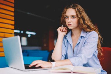 Ciddi bir kız öğrenci modern dizüstü bilgisayarla birlikte çalışma alanında oturuyor ve web sitesinden gelen bilgiler üzerinde kafa yoruyor, kıvırcık saçlı, netbook 'ta antivirüs güncellemesi bekleyen konsantre bir hippi kız.