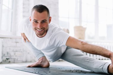 Neşeli beyaz erkek kameraya bakıyor kaslarını esnetiyor ve vücudunu evde çalıştırıyor. Dairesinde gülümseyen genç hippi, sağlıklı bir zihin ve beden için Asna 'da duruyor.