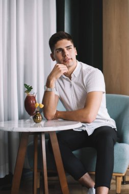 Genç bir erkek, gündelik kıyafetleriyle masada oturuyor dirseğine yaslanıyor ve rahat bir kafede dinlenirken yüzünü çeviriyor.